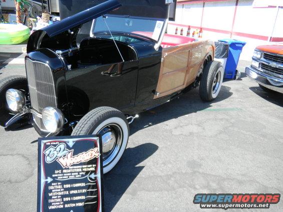 Black hot rod with wood paneling and red interior, hood open, parked next to a blue trash bin and an orange truck