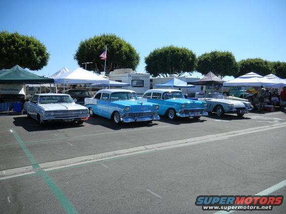 Several classic cars parked in a lot under tents, with trees and a clear blue sky in the background