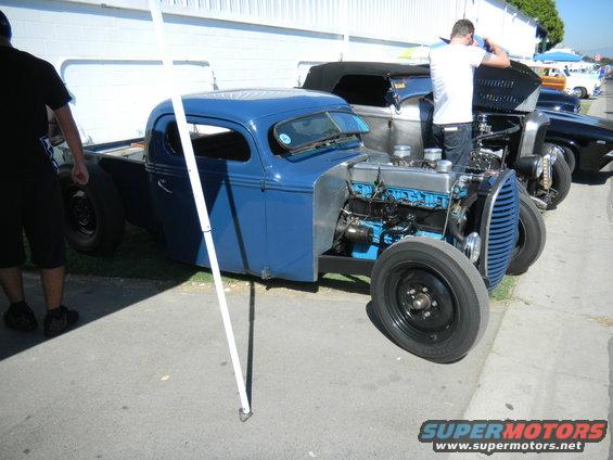 Blue hot rod truck with exposed engine and black wheels, parked outdoors