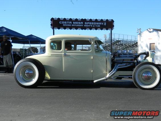 Custom hot rod coupe with exposed engine and whitewall tires parked near a speedway sign