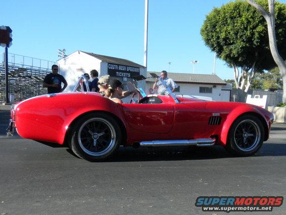 Red convertible sports car with side exhaust pipes and black wheels, seen from the side at a speedway