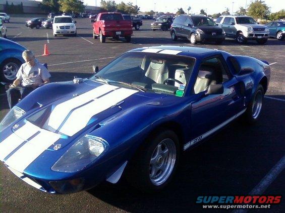 Blue Ford GT40 replica with white racing stripes parked in a lot, person sitting in a chair nearby