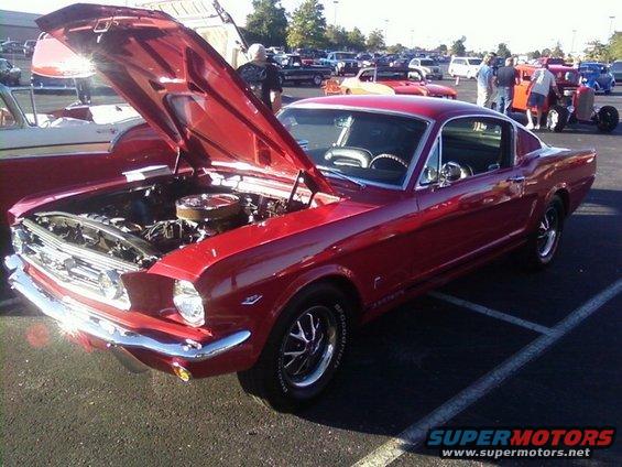 Red 1965 Ford Mustang Fastback with hood open, showing engine. Chrome bumpers and trim. BFGoodrich tires
