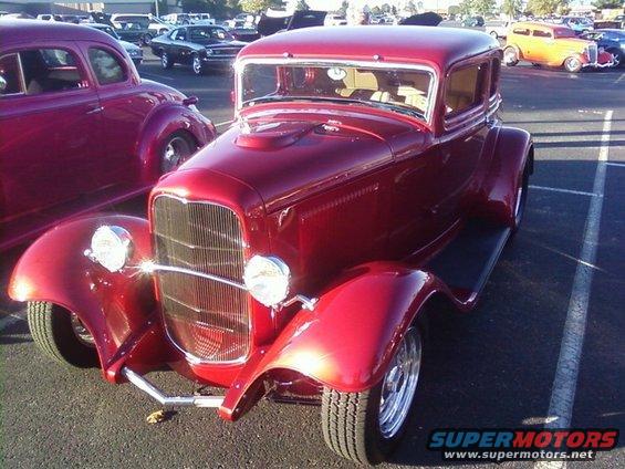 Red hot rod coupe parked in a lot with other cars. Front view shows grille and headlights