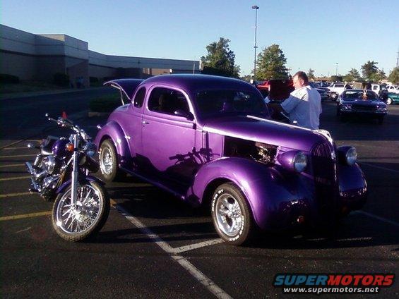 Purple vintage coupe with hood open parked next to a motorcycle. A man stands by the car