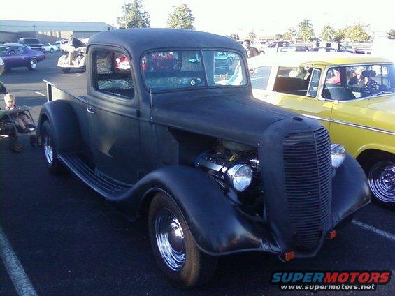 Black hot rod pickup truck with exposed engine and chrome wheels parked next to a yellow car
