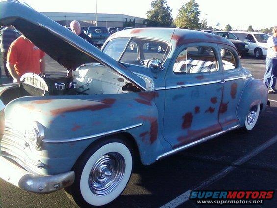 Blue sedan with primer patches, hood open, whitewall tires