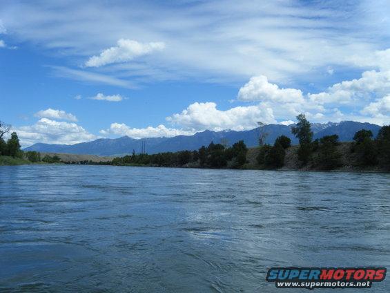 Wide river flows with mountains and trees in the background under a cloudy blue sky