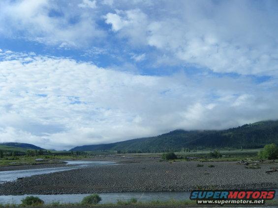 Wide riverbed with a shallow stream, leading to tree-covered mountains under a cloudy sky