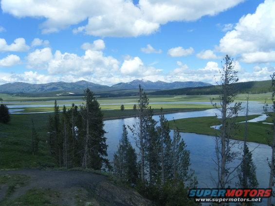 Landscape scene with a winding river, green fields, and distant mountains under a cloudy blue sky