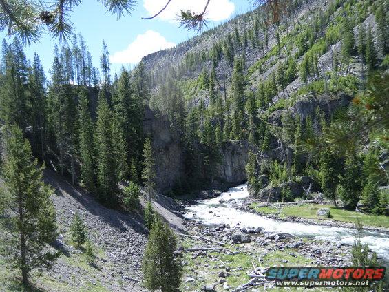 Mountain river valley with pine trees and rocky slopes under a blue sky. Watermark reads SUPER MOTORS