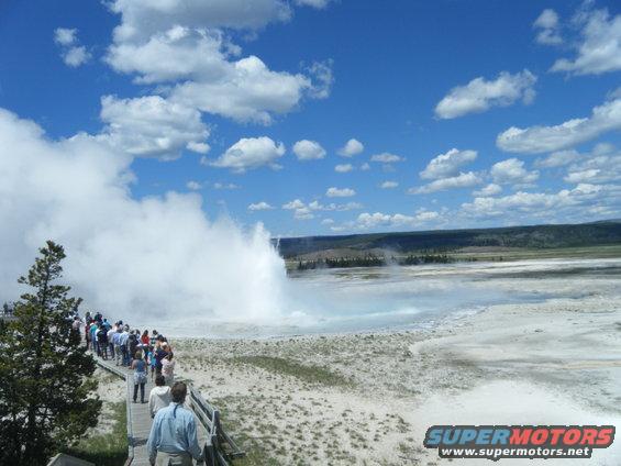 Wooden boardwalk with tourists watching a geyser erupt under a blue sky with white clouds