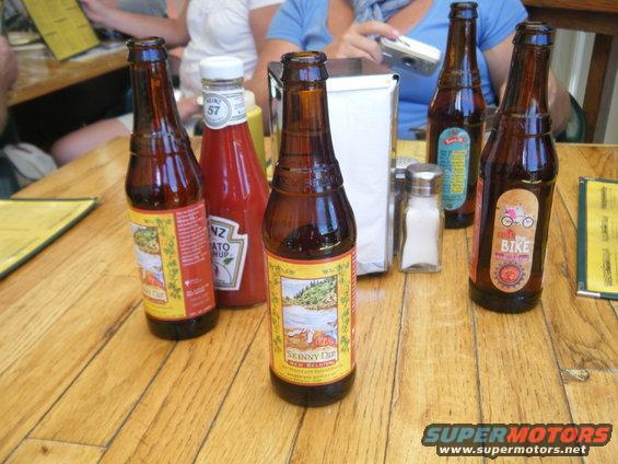 Several beer bottles and a Heinz ketchup bottle sit on a wooden table with people in the background