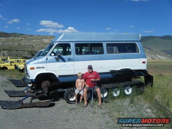 Man and kid next to a van with skis and snow tracks instead of wheels