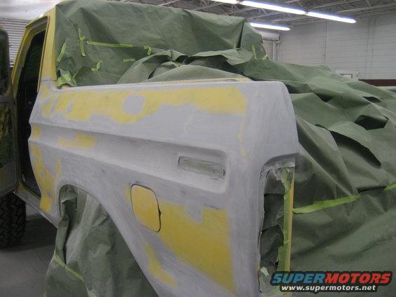 Gray and yellow primed Ford truck rear fender and bed corner, covered in green masking. Visible fuel door and taillight housing