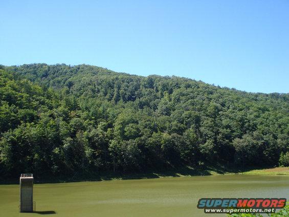 water reservoir with concrete structure and forested hill under clear blue sky. Supermotors.net logo at bottom right