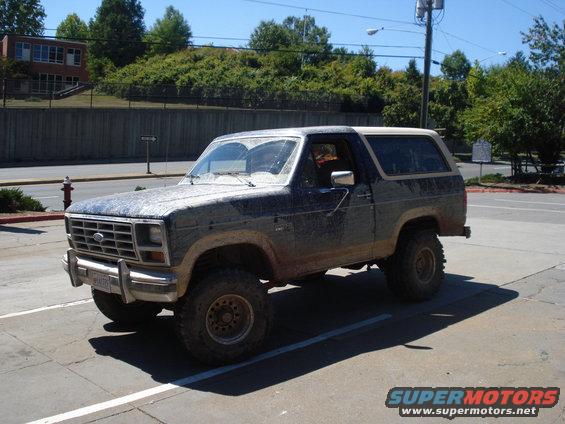 Mud-covered Ford Bronco parked on pavement. It has a dark body with a white top and off-road tires