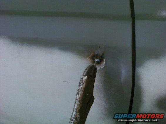 Drill bit creating a hole in a light-colored car panel, with debris flying