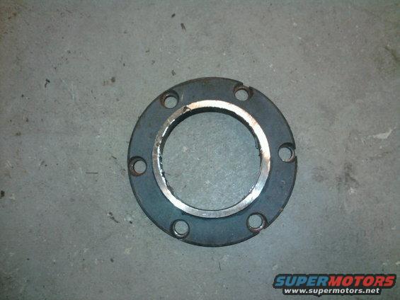 Dark metal flange with six bolt holes and a silver inner ring, lying on a concrete surface