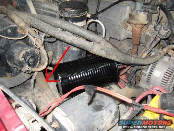 Close-up of engine bay with hoses, wires, and a black cylindrical part highlighted by a red arrow