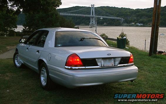 Silver 2000 Ford Crown Victoria parked on grass near a body of water, with a bridge in the background