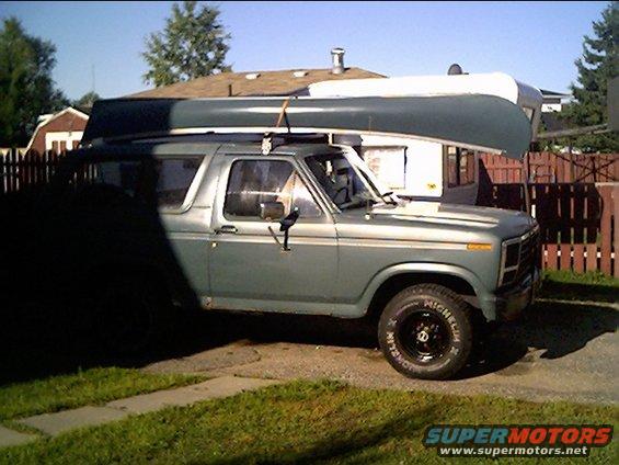 Side view of a light blue 1980s Ford Bronco with a canoe on the roof rack and a trailer behind it