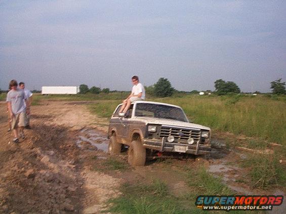 Brown and white Ford Bronco stuck in mud, with a person sitting on the roof