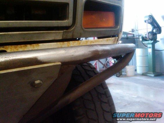 Close-up of a custom metal bumper with visible rust, attached to a vehicle's front end. An orange turn signal is visible above the bumper
