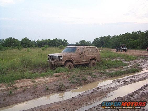 Mud-covered 1986 Ford Bronco navigating a muddy off-road course with another vehicle in the background