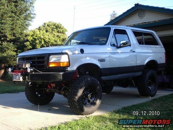 Lifted silver Ford Bronco parked in driveway, front 3/4 view. Has aftermarket bumper and lights