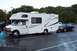 2005 Chevrolet Silverado 3500 - A white RV is parked on a wet lot, towing a dark sedan behind it.