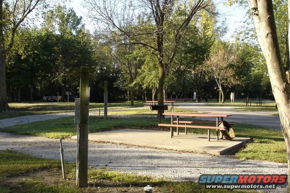 Campsite with picnic table, grill, and trees. A vehicle is barely visible in the background