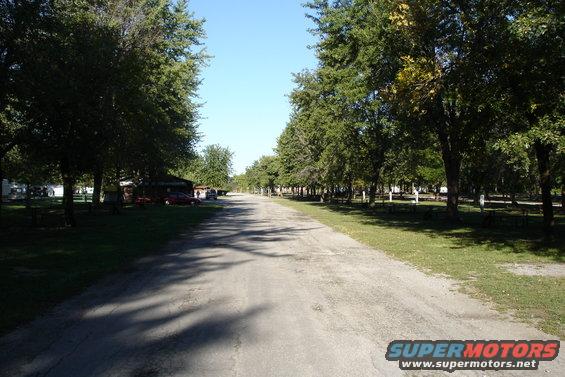 Dirt road through a campground with trees and picnic tables. A few cars are parked