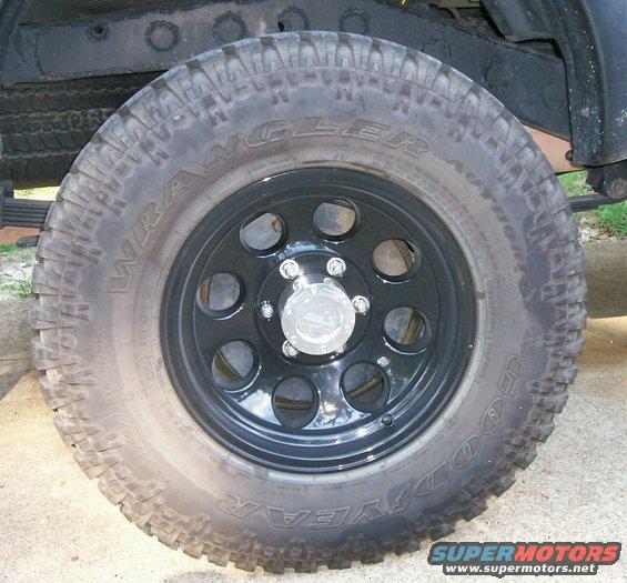Close-up of a black aftermarket wheel with Goodyear Wrangler tires. Hub cap has a logo