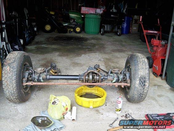 Front axle assembly with tires, differential visible, resting on concrete floor with tools and oil pan