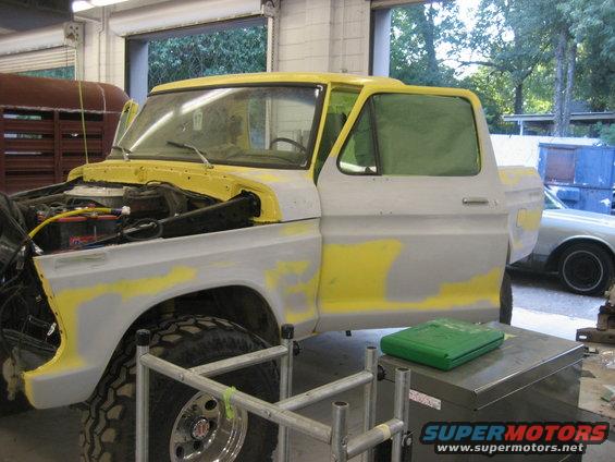 Ford Bronco body being prepped for paint, hood open, driver's door ajar. Another car is visible in the background