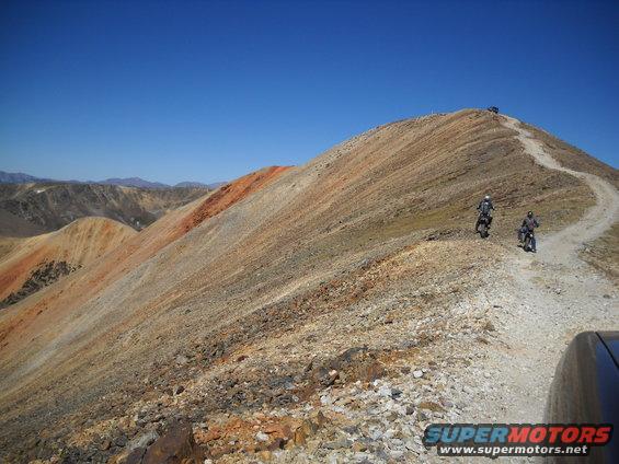 Two motorcycles ride up a dirt road on a mountain pass under a clear blue sky. A vehicle is visible at the top of the pass