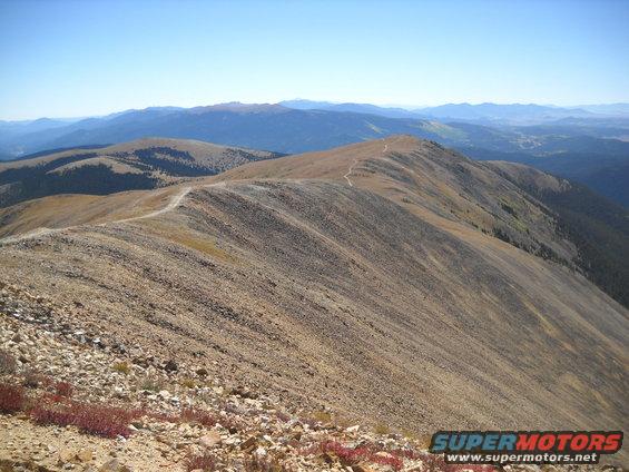 Mountain range with a winding dirt road on the ridge under a clear blue sky. Some low red plants in the foreground