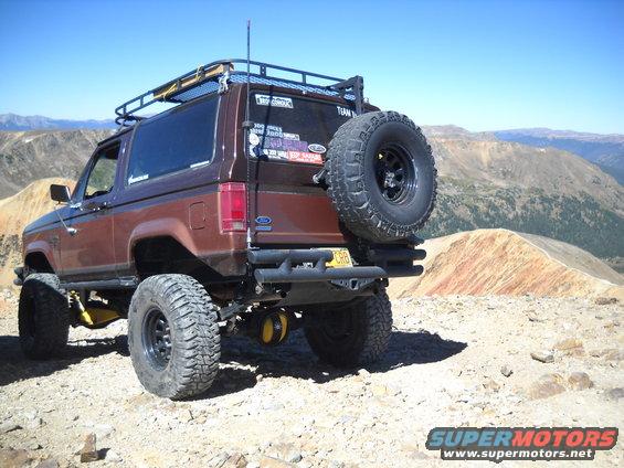 1986 Ford Bronco II parked on a rocky mountain trail, equipped with a roof rack and spare tire carrier