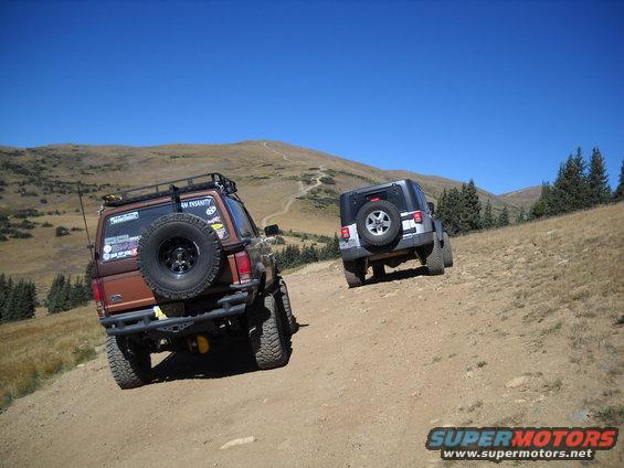Brown SUV with roof rack and spare tire on rear bumper climbs dirt hill, followed by a silver Jeep. Mountains and blue sky in background