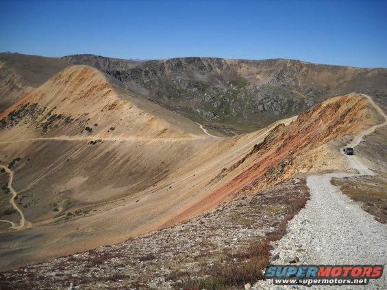 Vehicle on a dirt road winding through a mountainous landscape under a clear blue sky