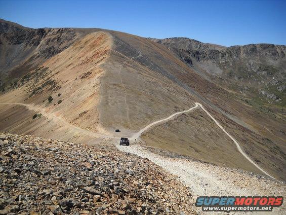 Two SUVs driving on a winding dirt road up a steep mountain pass under a clear blue sky