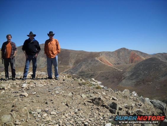Three men in cowboy hats stand on a rocky mountain peak overlooking a vast, arid landscape with winding roads