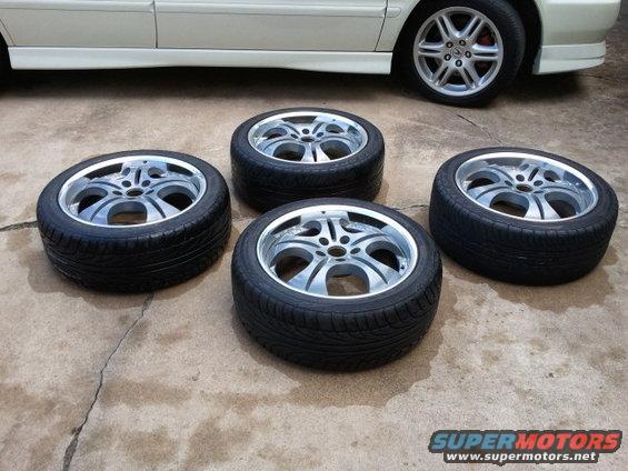 four shiny aftermarket wheels with tires on concrete, part of a white car visible in background