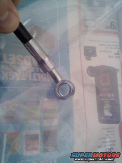 Metal brake line fitting with a banjo bolt hole, held by a thumb and forefinger. The background shows a blurry catalog page with product images and text