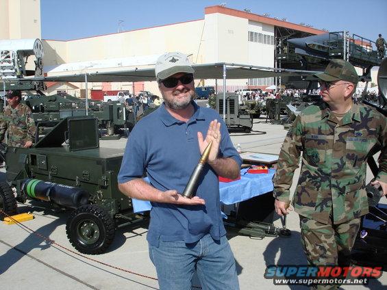 Man holding a large bullet with military equipment in background