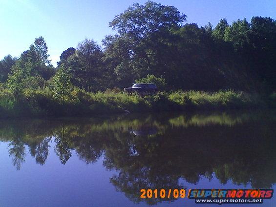 Dark SUV parked on grassy bank beside a lake, reflected in water