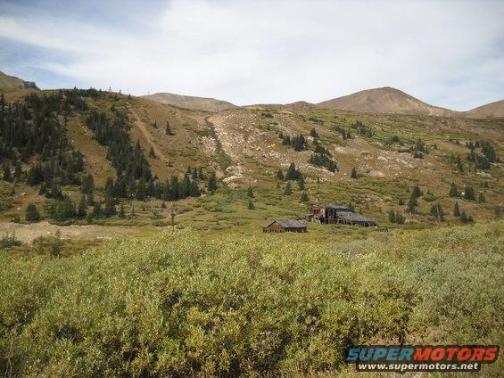 Abandoned buildings sit in a grassy valley below a rocky mountain slope with scattered trees