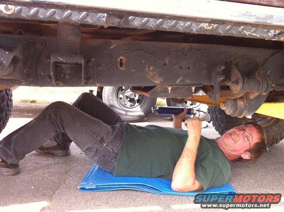 Man on blue mat working on vehicle frame with grease gun. Another vehicle's wheel is visible