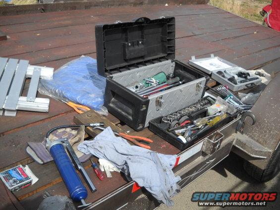 Toolboxes and various tools spread out on a wooden trailer deck. Includes a blue grease gun, wrenches, sockets, and pliers